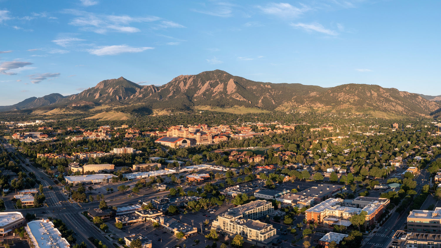 Aerial view of a vibrant city with a sprawling university campus, nestled amidst lush greenery and majestic mountains under a clear blue sky.